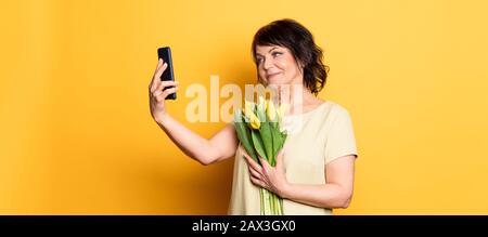 Belle vieille femme avec maquillage professionnel fumé et coiffure tenant des fleurs de tulipe sur le fond rose. Prise d'un appel vidéo par le smartphone Banque D'Images