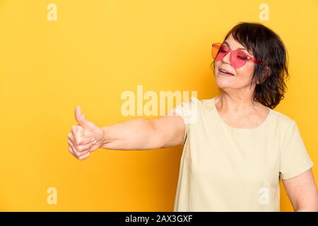 Belle femme Senior en rose néon lunettes de soleil montrant comme par doigts sur fond jaune Banque D'Images