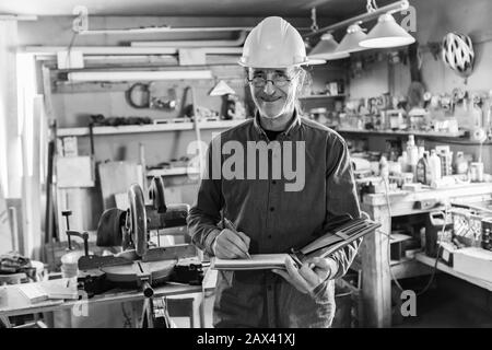 Inspecteur dans le garage pendant une inspection. Homme du caucase d'âge moyen portant un casque en souriant et écrit des notes sur un ordinateur portable, noir et blanc Banque D'Images