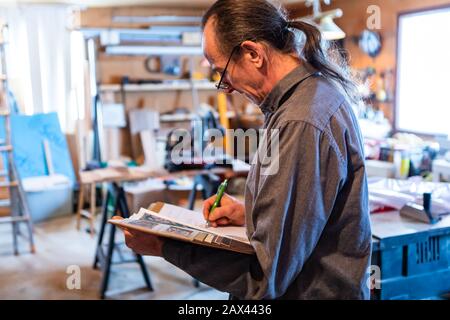 homme d'âge moyen avec de longs cheveux portant des lunettes dans le garage avec des outils en arrière-plan. inspecteur à la maison prenant des notes professionnelles sur son ordinateur portable Banque D'Images