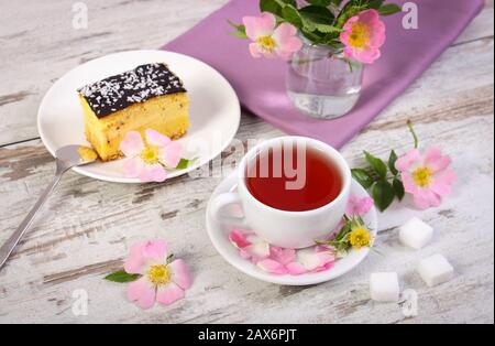 Tasse de thé chaud avec rose sauvage, fleurs fleuries et cheesecake sur plaque sur fond rustique en bois vieux Banque D'Images