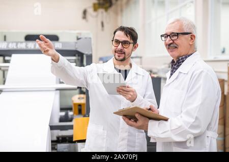 Un expert souriant de qualité barbu dans les verres gestant la main et utilisant la tablette tout en discutant de la productivité de l'équipement de maison d'impression avec un collègue Banque D'Images