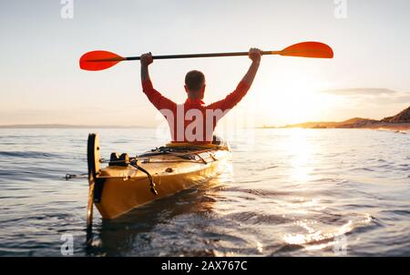 La vue arrière de l'homme maintient les palettes de kayak en hauteur. Senior actif dans un kayak sur la mer du coucher du soleil Banque D'Images