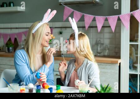 Portrait de la mère drôle et de la fille pendant la préparation pour Pâques. Ils portent des oreilles de lapin. Mère peinture du nez de fille. Banque D'Images