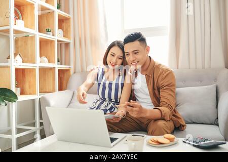 Smiling young couple s'asseoir sur la table à l'aide d'ordinateur portable en prenant soin de factures de services publics et de la chambre, lire la paperasse Banque D'Images