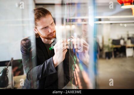 Jeune homme travaillant avec des bases de données et des diagrammes, écrivant des idées sur le mur de verre de bureau Banque D'Images