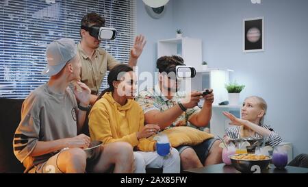 Deux jeunes hommes jouant au jeu en ligne dans des lunettes de réalité virtuelle, un gars gagne la bataille. Leurs amis les encouragent, mangeant des collations et buvant des cocktails de glace sèche. Banque D'Images