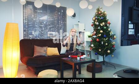 La jeune femme passe des appels vidéo par smartphone avec des gratte-ciel en arrière-plan. Elle est assise sur un canapé avec un verre de boisson dans le salon décoré avec un arbre de noël et des lumières parmi la fumée. Banque D'Images