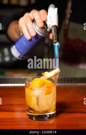 Un barman mâle brûle un bâton de cannelle avec un brûleur à gaz dans un bar. Processus de préparation d'un cocktail. Banque D'Images