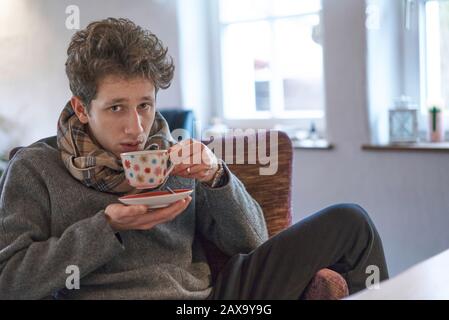 le jeune homme est assis sur le fauteuil à la maison et boit une tasse de thé donnant un peu malade Banque D'Images