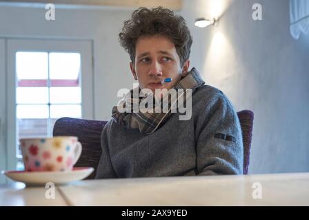 le nuage de jeune homme se trouve à la table à la maison avec un foulard autour de son cou et a un thermomètre dans sa bouche, il semble comme s'il a un froid Banque D'Images