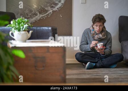 Jeune homme beau avec un froid assis sur le sol dans son appartement avec un thermomètre dans sa bouche et une tasse de thé dans ses mains, regardant déprimé Banque D'Images