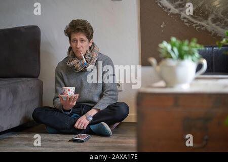 Jeune homme déprimé avec un froid assis sur le sol dans son appartement avec un thermomètre dans sa bouche et une tasse de thé dans sa main Banque D'Images