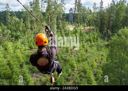 Cavalier mâle avec casque orange sur la ligne Little Rock Zip au-dessus d'une forêt de pins sur le chemin d'une tour d'atterrissage en bois au loin Banque D'Images