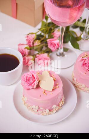 Mini gâteau avec glacer rose, belles roses, tasse de café, verre de vin rose sur la table blanche Banque D'Images
