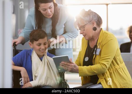 Médecin avec tablette numérique parlant au jeune patient en fauteuil roulant et mère dans le hall de la clinique Banque D'Images