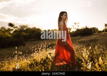 Belle jeune fille avec des cheveux bouclés en robe orange vif promenades à travers la forêt de l'été ou d'automne ou sur le terrain au coucher du soleil. Femme est souriante et ha Banque D'Images