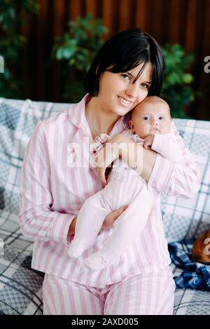 Une mère en pyjama est assise sur un canapé et tient un bébé dans ses bras. Banque D'Images