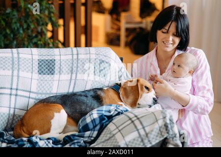 Une mère en pyjama tient un bébé dans ses bras à côté d'un chien allongé sur le canapé. Banque D'Images