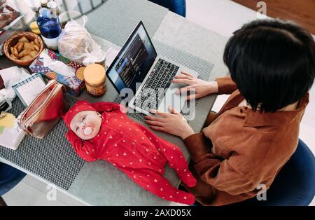 La mère est assise et travaille sur un ordinateur portable près du bébé allongé sur la table. Concept d'emploi des femmes avec des bébés. Vue de dessus. Banque D'Images