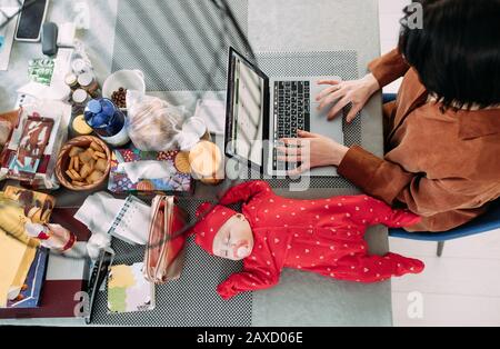 La mère est assise et travaille sur un ordinateur portable à côté de dormir bébé sur la table. Concept d'emploi des femmes avec des bébés. Vue de dessus. Banque D'Images