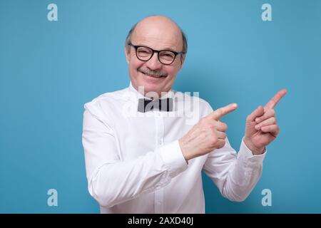Bon homme senior regardant avec l'expression satisfaite, indique avec les doigts avant de côté sur le copyspace, vous montre l'endroit intéressant. Concept de publicité Banque D'Images