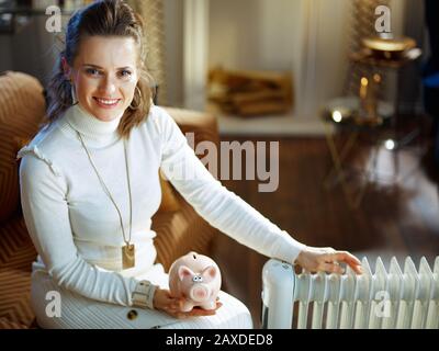 une femme de ménage souriante et tendance dans un pull blanc et une jupe dans la maison moderne en hiver ensoleillé assis sur un canapé près de l'huile électrique blanche radiat Banque D'Images