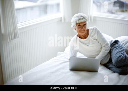 Femme mûre dans son pyjama se trouve sur un lit penchant sur son coude et regarde d'un ordinateur portable comme elle pose pour un portrait. Banque D'Images