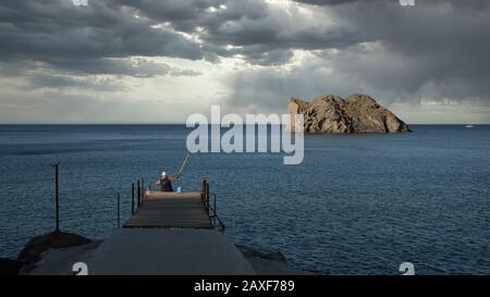 Pêcheur assis sur un quai délabré lors d'une journée de tempête avec l'île en arrière-plan sur l'île de Lesvos en Grèce Banque D'Images