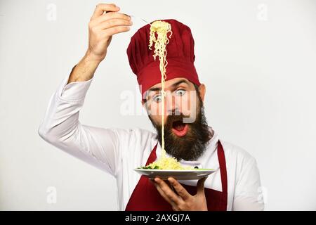 Cuire avec un visage choqué en bordeaux uniforme maintient la fourche et la plaque. L'homme avec barbe conserve un plat savoureux sur fond blanc. Le chef mange des nouilles italiennes ou asiatiques. Concept de restauration rapide. Banque D'Images