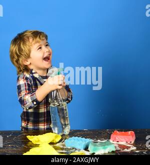 Enfant près de la table avec des fournitures de nettoyage. Garçon en chemise à carreaux sur fond bleu. Enfant en riant le visage vaporisant de l'eau hors de la bouteille de pulvérisation. Concept d'activités de nettoyage. Banque D'Images