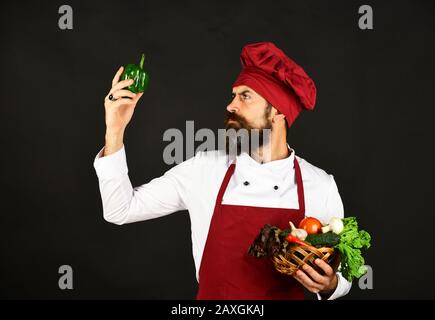 Homme avec barbe sur fond noir regarde le poivre. Le chef tient de la laitue, de la tomate, du poivre et des champignons. Cuisinez avec un visage sérieux en bordeaux uniforme contient des légumes dans un bol en osier. Concept d'épicerie verte Banque D'Images