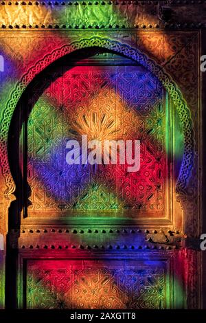 Porte traditionnelle, ancienne et marocaine avec sculptures en bois, avec des reflets multicolores de la fenêtre en verre colorée dans le palais de Bahia à Marrakech, Maroc. Banque D'Images