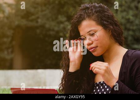 Jolie femme latine qui ajuste les lunettes tout en travaillant sur un ordinateur portable sur le campus de l'université de parc. Portrait avec espace de copie pour le texte Banque D'Images