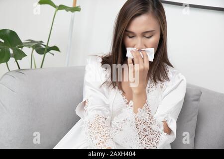 Une femme asiatique dans une gigole en satin et un peignoir de dentelle blanche a eu la grippe, assis sur un canapé gris dans le salon. La fille en dentelle vêtements de nuit a un snot at Banque D'Images