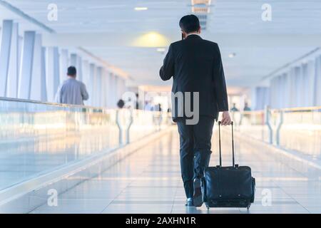 Businessman walking et roulant un chariot valise dans le hall, parlant sur un téléphone mobile. Concept de voyages d'affaires. Banque D'Images