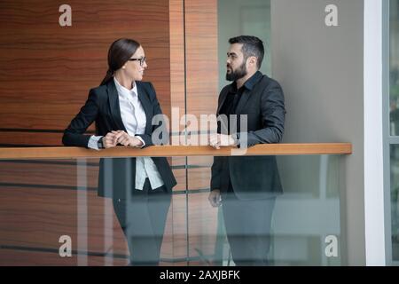 Homme barbu en costume debout sur le balcon au bureau et parler à une collègue de femme dans des lunettes Banque D'Images