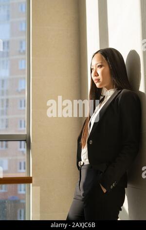 Jeune femme grave pensive asiatique en costume noir debout au mur dans le couloir Banque D'Images