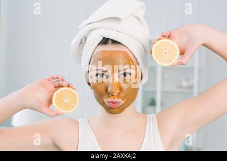 Jolie fille avec masque facial marron tenant une tranche de citron près de son visage et souriant. Photo de fille recevant des soins de spa. Beauté et soins De La Peau Banque D'Images