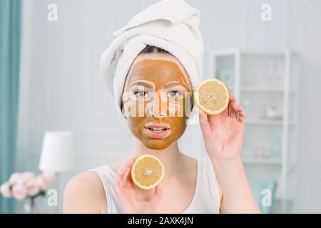 Jolie fille avec masque facial marron tenant une tranche de citron près de son visage et souriant. Photo de fille recevant des soins de spa. Beauté et soins De La Peau Banque D'Images