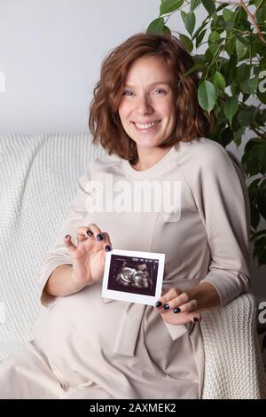 Portrait d'une femme enceinte souriante en robe beige tenant une échographie, montrant un sonogramme de bébé tout en étant assis sur un canapé à la maison, ve Banque D'Images