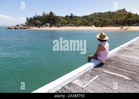 Homme de cheveux bouclés, avec chapeau de soleil, assis sur le bord du quai en bois Banque D'Images