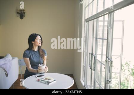 Une jeune femme réfléchie qui a du café tout en regardant par la fenêtre dans le salon à la maison Banque D'Images