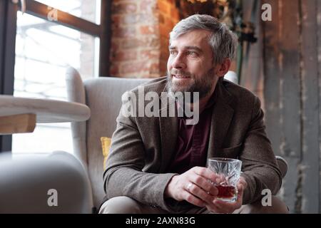 Homme à la barbe de rêve d'âge moyen dans une veste marron assise dans un fauteuil et buvant de l'alcool Banque D'Images