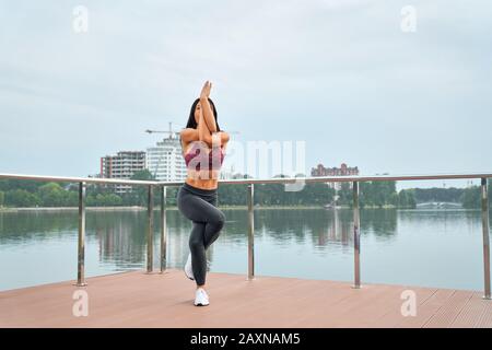 Portrait complet d'une femme en bonne santé avec un corps fort portant une tenue de sport debout sur un pied tout en tordant les jambes et les bras près du lac de la ville. Jolie dame pratiquant le yoga à l'extérieur. Banque D'Images