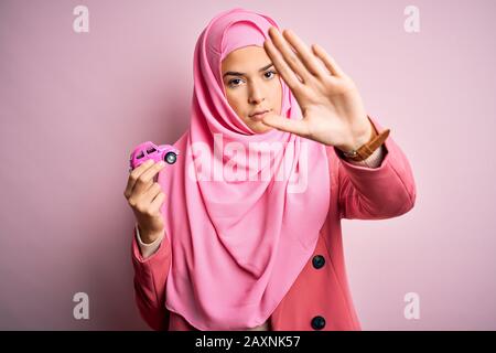 Jeune fille portant un hijab musulman tenant un petit jouet de voiture sur un fond rose isolé avec main ouverte faisant signe d'arrêt avec expressi sérieux et confiant Banque D'Images