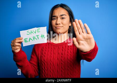 Jeune femme asiatique belle tenant du papier demandant de sauver la terre et la conservation de l'environnement avec main ouverte faisant signe d'arrêt avec sérieux et confiant Banque D'Images