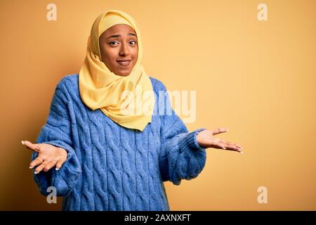 La jeune afro-américaine africaine porte le hijab musulman sur un fond jaune isolé sans indice et l'expression confuse avec les bras et les mains soulevées. Fais Banque D'Images