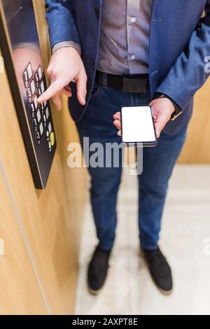 Mains de l'homme tenant un téléphone portable à écran vide lors de l'utilisation du panneau de commande de l'élévateur. Banque D'Images