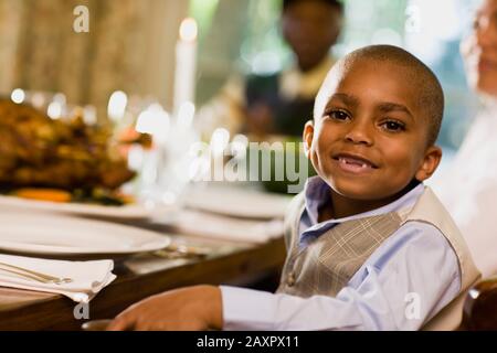 Jeune garçon sourit comme il pose pour un portrait tout en étant assis à une table de salle à manger avec une bougie et de la nourriture avec une femme et un homme en arrière-plan. Banque D'Images